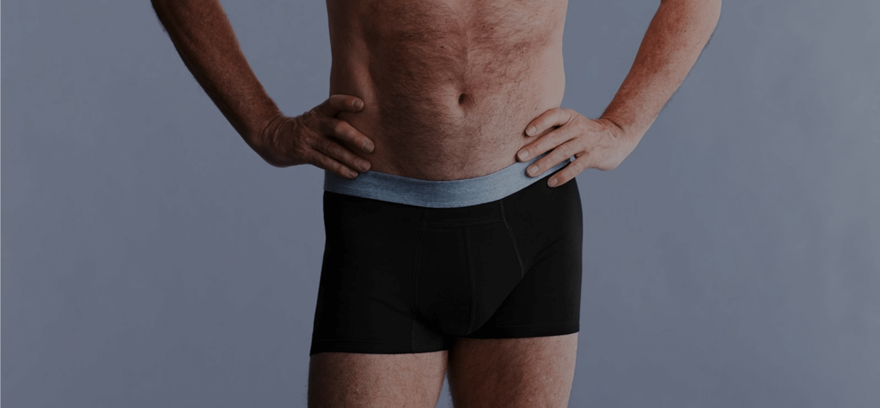 Mann steht mit den Händen in der Hüfte, trägt schwarze schützende TENA Men Inkontinenz-Boxershorts vor neutralem grauen Hintergrund.