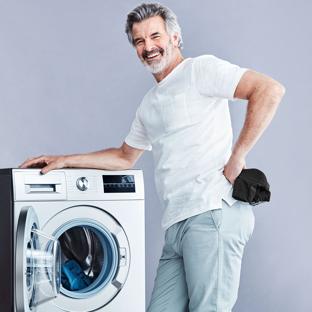 Ein Mann hält ein Paar schwarze Herren-Inkontinenzboxershorts in der Hand, lächelt und lehnt sich auf eine Frontlader-Waschmaschine vor grauen Hintergrund.